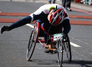 Zdjęcie przedstawia sportowca w kasku na wyczynowym wózku inwalidzkim niorącego udział w wyścigu.