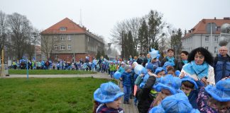 Zdjęcie przedstawia dzieci z niebieskim kapelusikami maszerujące chodnikiem, w oddali widać, że pochód jest długi, zakręca i kończy się poza kadrem.