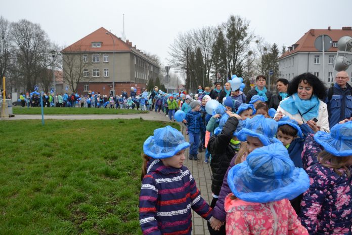 Zdjęcie przedstawia dzieci z niebieskim kapelusikami maszerujące chodnikiem, w oddali widać, że pochód jest długi, zakręca i kończy się poza kadrem.