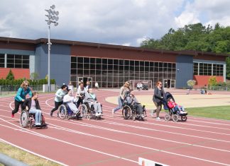 Zdjęcie: w tle hala sportowa, na pierwszym planie, na bieżni pięć osón na wózkach iwalidzkich pędzi wzdłuż torów - pomagają im opiekunki popychające wózki.