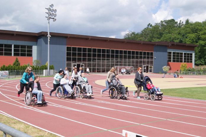 Zdjęcie: w tle hala sportowa, na pierwszym planie, na bieżni pięć osón na wózkach iwalidzkich pędzi wzdłuż torów - pomagają im opiekunki popychające wózki.